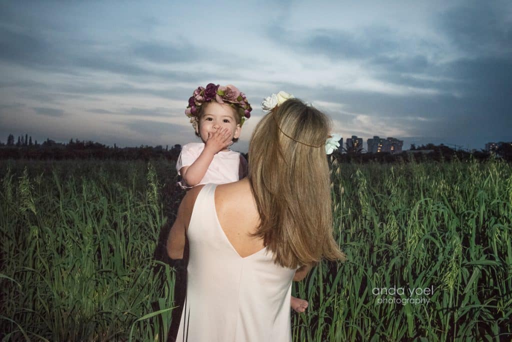 ליהיא גרינר בגבה למצלמה מחזיקה את בתה בידיים בשדה חיטה ירוק בשקיעה - מסדרת צילומי המשפחה בטבע אנדה יואל